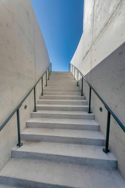 Austin, Texas- Staircase with concrete steps in the middle of concrete walls. Stairs with floor-mounted metal handrails near the walls and a view of a clear blue sky background.
