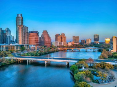 Günbatımında Austin, Teksas şehir manzarası yakınlarındaki Colorado Nehri üzerindeki park ve köprüler. Parkta sağda insanlar var ve gökyüzü ufkunda gün batımı parlayan binalar var..