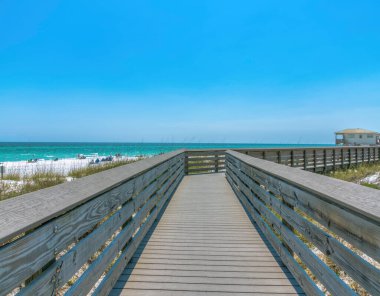 Destin, Florida 'da insanlarla birlikte sahilde tahta kaldırım. Kenarlarında çimen manzaralı sahil yolu ve sağında mavi gökyüzüne karşı mavi su ve sahil evi manzaralı..