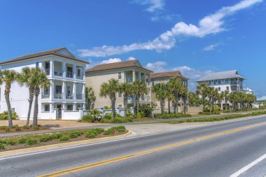 Destin, Florida 'da bir plajın yakınında balkonları ve palmiye ağaçları olan üç katlı sahil evleri. Önünde otoban bulunan kaldırımlı evler ve mavi gökyüzü arkaplanı manzarası..