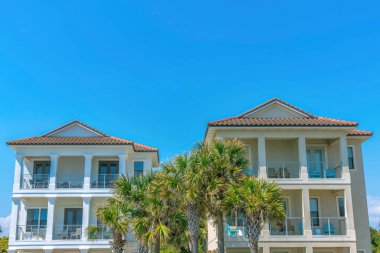 Destin, Florida. Ortasında palmiye ağaçları olan iki sahil evi. Mavi gökyüzü arka planına karşı balkonları olan yazlık evlerin dışı..