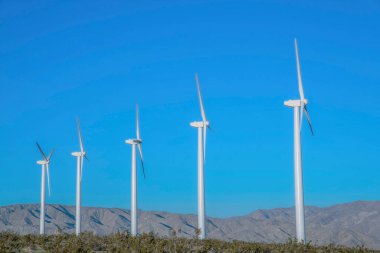 Kaliforniya 'daki dağ sırasına ve mavi gökyüzüne karşı beyaz borulu rüzgar türbinleri. Dağ sırasının yakınındaki bir çöl çiftliğinde rüzgar türbinleri..
