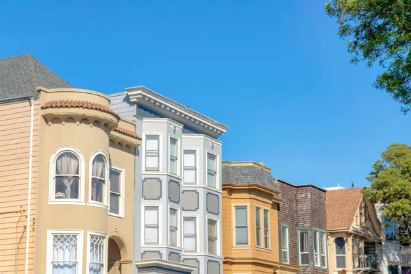 Row of houses with different structures against the clear blue sky in San Francisco, California. Neighborhood houses exterior with curved walls, bow windows, and shingles sidings and a view of trees.