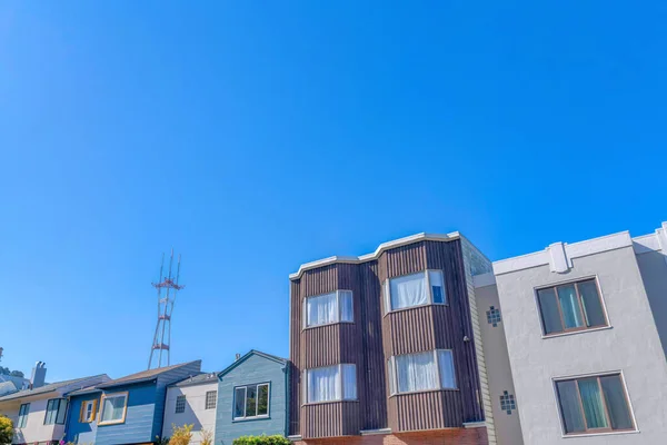 Adjacent suburban houses against the Sutro Tower and clear blue sky background at San Francisco, CA. There is a house on the right with light gray exterior along with the houses with wood sidings.