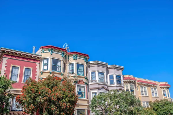 Colorful apartment flat buildings at San Francisco, California. Adjacent residential buildings with emergency stairs and trees at the front and a clear blue sky at the back.