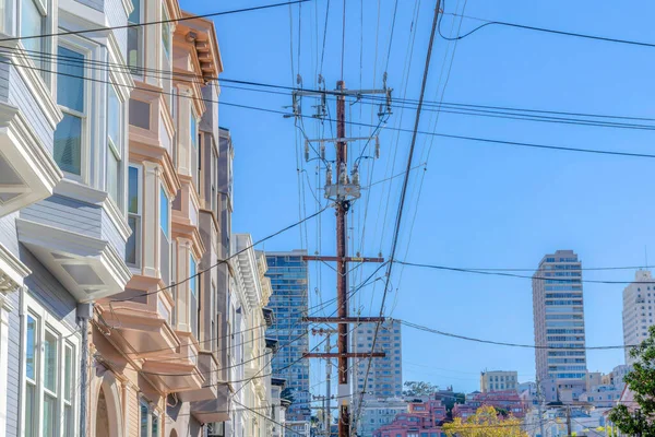 Intersecting electrical wires and electric posts in an urban area at San Francisco, CA. There is a row of victorian townhomes on the left beside the posts against the high-rise buildings at the back.