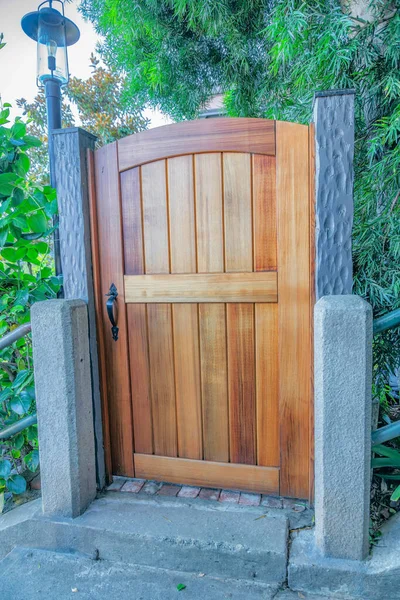 Wooden single gate entrance with concrete pillars and lamp post- San Francisco, California. Entrance with wooden gate and black door handle against the plants and trees at the back.