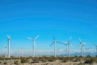 Dağları olan yel değirmenleri ve güneşli bir günde açık mavi gökyüzü. Güneşli bir alanda üç pervaneli rüzgar pompaları alternatif enerji üretiyor..