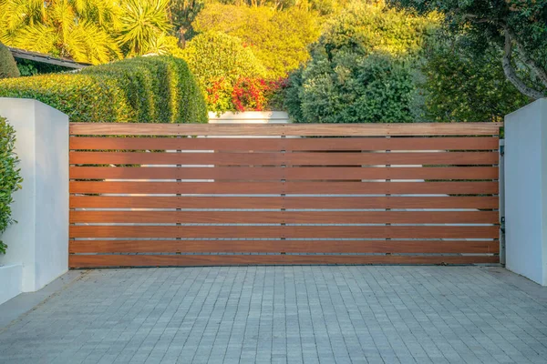 La Jolla, California- Wide wooden gate of a residence. There is a concrete pavement at the front of the gate against the bushes and trees at the back.