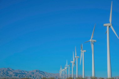 Rüzgâr türbinleri ve Kaliforniya 'da bir çölde çelik boru kulesi. Dağ sırasına ve mavi gökyüzüne karşı beyaz yel değirmenleri var..