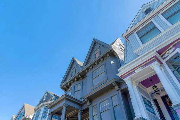 View of victorian houses from below against the sky in San Francisco, CA. There is a gray house on the left with decorative porch pillars and bay windows beside the house with decorative trims.