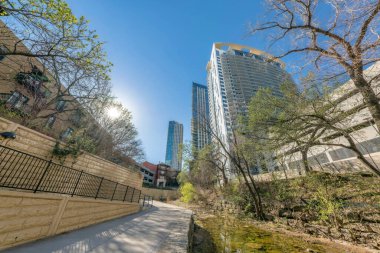 Austin, Teksas... Nehir kanalı yakınlarındaki ağaçlar ve gökdelenler manzaralı yürüyüş yolu. Solda bir bina duvarının yanında bir yol ve sağdaki modern binaların manzarası var..