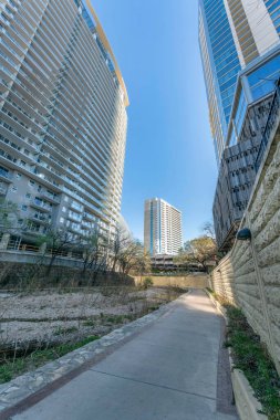 Austin şehir merkezi, Teksas. Modern binaların ortasında nehir kanalının yanındaki patika. Solda balkonlu ve sağında sarı bir duvar olan bir bina var..