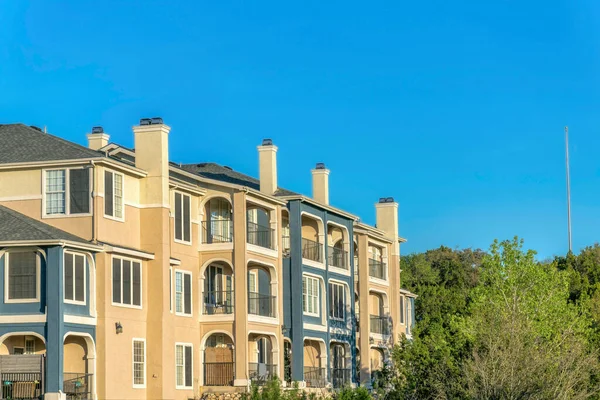 Austin, Texas- Apartment building near Lake Austin with blue and beige exterior. Residential building exterior with balconies and a view of trees on the right.