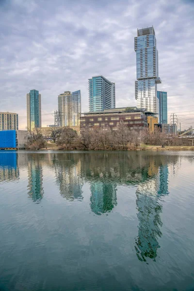 Colorado River waterfront with a reflection of the cityscape at Austin, Texas. There are trees at the front of modern high rise buildings against the cloudy sky.