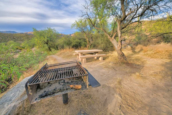 In ground wood and charcoal grill on a camping ground at Sabino Canyon in Tucson, Arizona. Grill near a concrete table and chairs against the desert plants and mountain at the back.