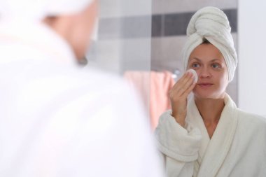 Bornozlu ve kafasında havlu olan genç bir kadın aynanın önünde kozmetik ürünlerini yıkıyor.