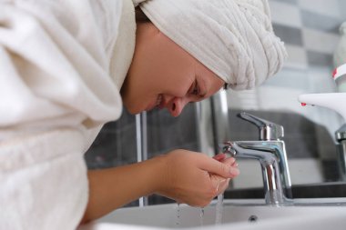 Bornoz ve havlu giymiş genç bir kadın yüzünü suyla yıkıyor.