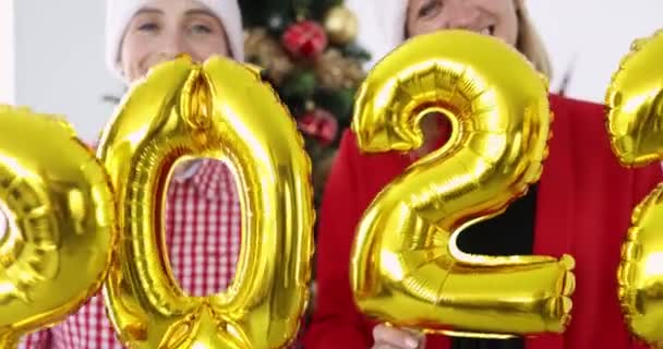 Молоді жінки в капелюхах Санта Клауса стоять біля ялинки і тримають повітряні гелеві кульки з номерами 2022 4k фільм — стокове відео