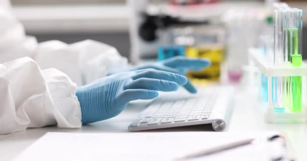 Vědec chemik v ochranném obleku a rukavice psaní na klávesnici v blízkosti zkumavky v laboratoři 4k film — Stock video