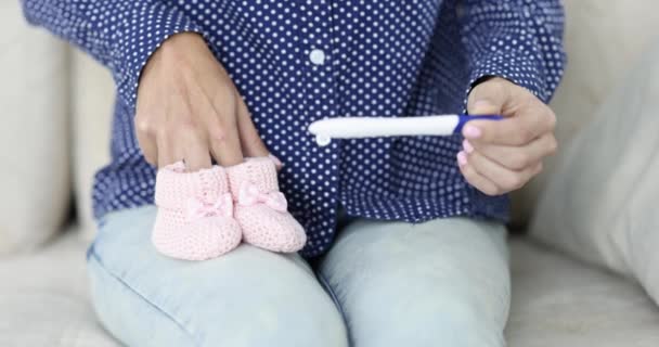 Mujer sentada en sofá con prueba de embarazo y sosteniendo botines rosados 4k película — Vídeo de stock
