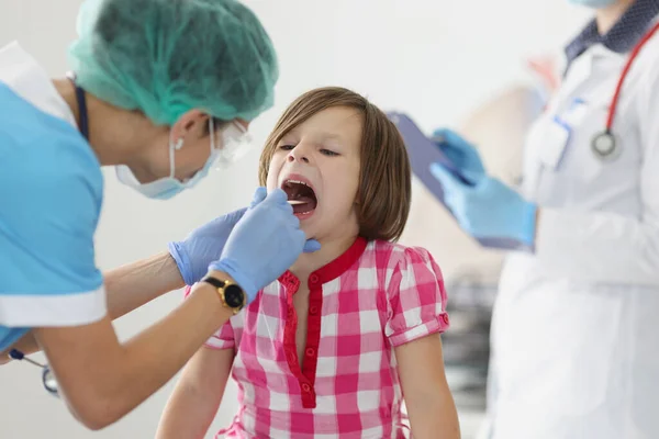 Otorhinolaryngologue médecin en masque médical de protection examinant la gorge de la petite fille en utilisant une spatule en clinique — Photo