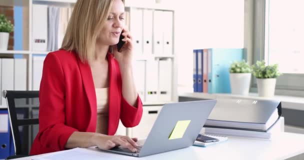 Молодая женщина в красной куртке говорит по телефону и печатает на клавиатуре компьютера 8k — стоковое видео