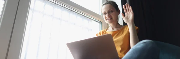 Jovem mulher acenando bem-vindo no laptop enquanto sentado no peitoril da janela — Fotografia de Stock