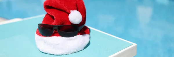 Chapeau santa claus rouge et lunettes de soleil couché sur la chaise longue sur le rivage de la piscine gros plan — Photo