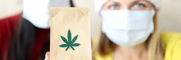 Kvinnor i röd tomte klo hattar och skyddande medicinska masker hålla papperspåse med marijuana extrakt närbild — Stockfoto