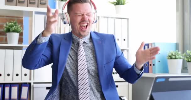 Joven hombre de negocios baila felizmente en los auriculares delante del monitor de la tableta 4k película — Vídeo de stock