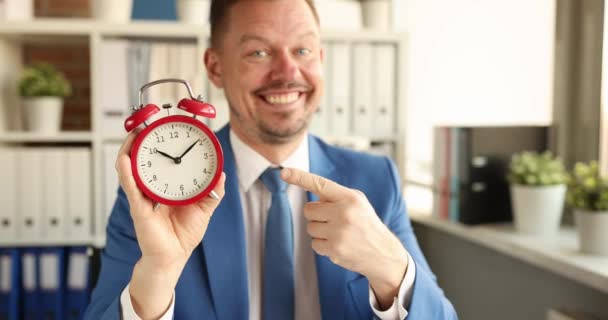 Junger lächelnder Geschäftsmann zeigt mit dem Zeigefinger die Zeit auf dem Wecker — Stockvideo