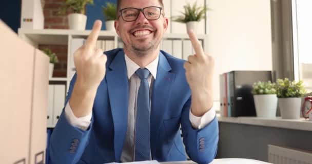 Joven hombre de negocios sonriente sentado en la mesa y mostrando el dedo medio 4k película — Vídeos de Stock