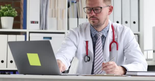 年轻医生，戴着眼镜，凝视着手提电脑屏幕，在医疗文件4k中写作 — 图库视频影像