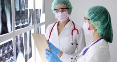 Koruyucu maskeli ve gözlüklü kadın doktor tıbbi belgelere ve röntgenlere bakıyor.