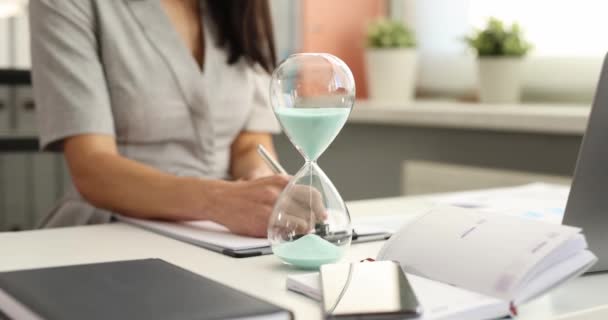 Blauw zand gieten in glas klok tegen de achtergrond van het bedrijfsleven vrouw werken aan tafel closeup 4k film slow motion — Stockvideo