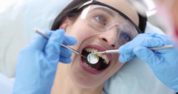 Стоматолог, осматривающий зубы пациентки в защитных очках с помощью металлических инструментов 4k фильм замедленной съемки — стоковое видео