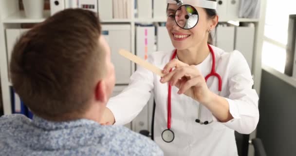 Врач с фронтальным зеркалом осматривает горло пациента деревянным лопатком 4k фильма замедленной съемки — стоковое видео
