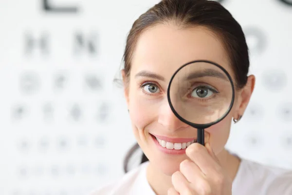 Mulher optometrista segurando lupa nos olhos contra fundo da tabela de exame de visão — Fotografia de Stock