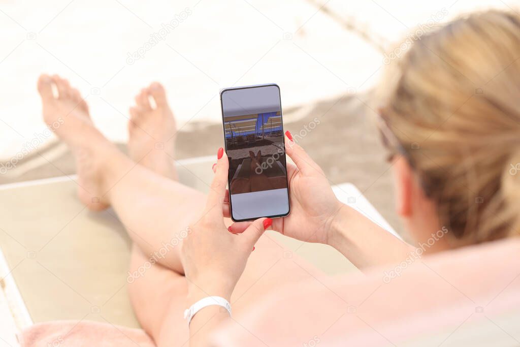 Young woman lying on sun lounger on beach and taking pictures of her feet on mobile phone closeup