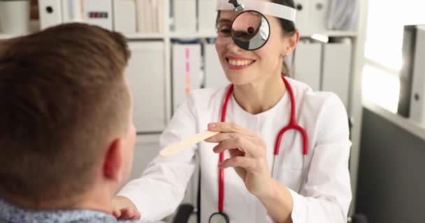 Лікар з лобовим рефлектором на лобі, який вивчає горло пацієнта чоловічої статі з шпателем 4k плівкою повільний рух — стокове відео