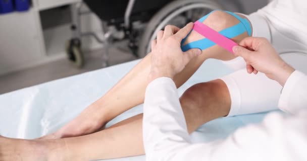 Doctor lijmen kinesiotape op patiënt zere knie in kliniek closeup 4k film slow motion — Stockvideo