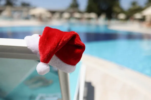 Chapéu de Papai Noel colocado na cadeira de gramado perto da piscina com água azul-turquesa limpa — Fotografia de Stock