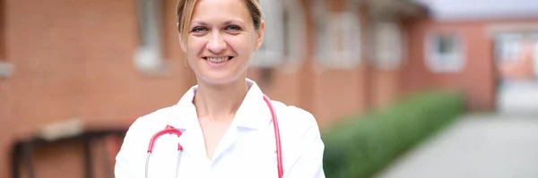 Портрет молодой улыбающейся женщины-врача с красным стетоскопом возле клиники — стоковое фото