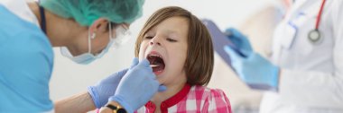 Korumacı tıbbi maskeli bir doktor, klinikte spatula kullanan küçük bir kızın boğazını inceliyor.