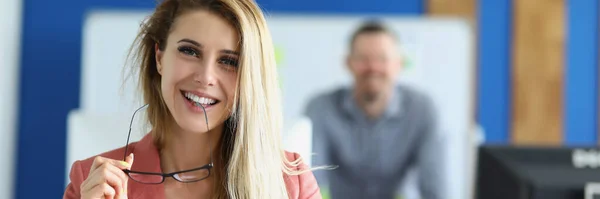 성공 한 젊은 여성은 입 가까이에 안경을 들고 사무실에서 동료들에 대항하여 웃고 있다 — 스톡 사진
