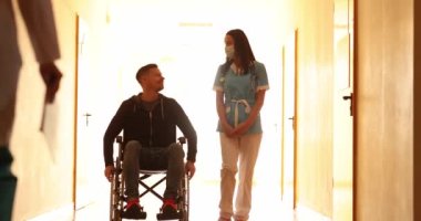 Hemşire tekerlekli sandalyeli hastayla koridorda yürüyor ve doktor 4K filmi ağır çekimde selamlıyor.