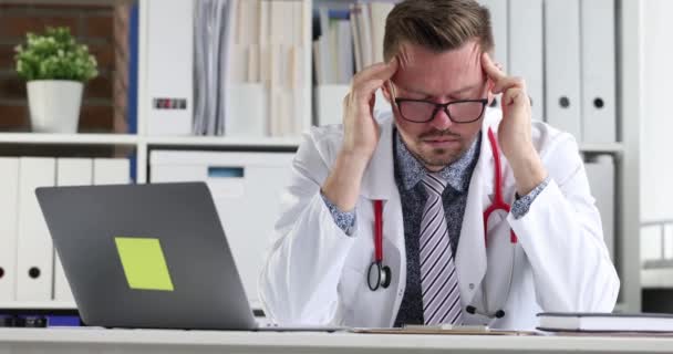 Cansado médico sentado delante de la computadora portátil y masajeando su templo 4k película cámara lenta — Vídeo de stock