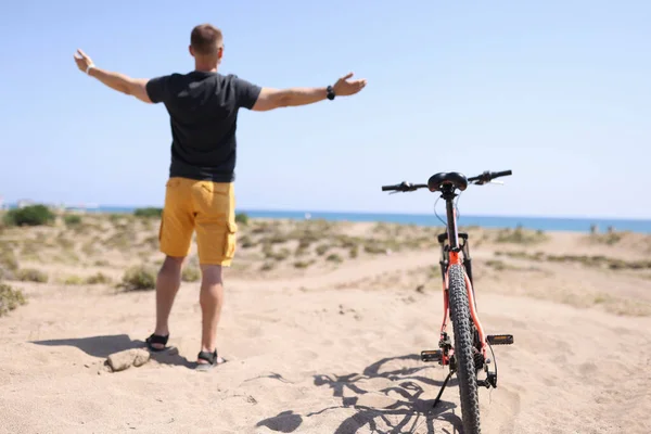 Uomo in piedi sulla spiaggia vicino alla bicicletta con le braccia divaricate vista posteriore — Foto Stock