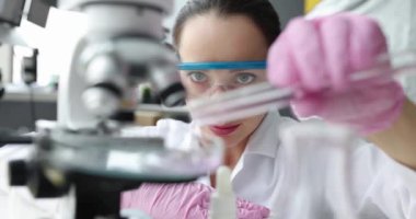 Kadın kimyager mikroskop önünde deney tüpünden petri kabına sıvı döküyor.